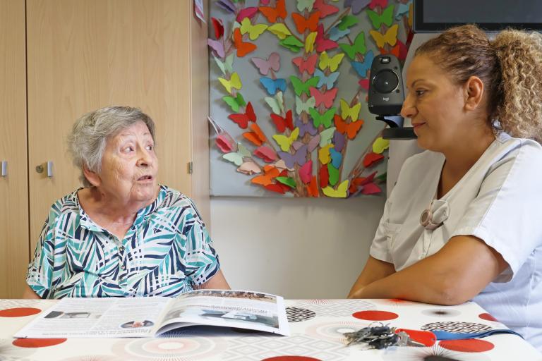 Hermine, résidente en EHPAD, est accompagnée d'une aide-soignante pour échanger autour d'une table.